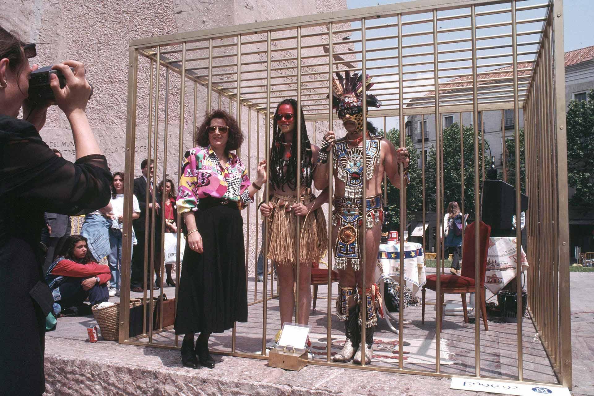 Three people stand inside a large golden cage outdoors. One wears modern clothes, another wears a grass skirt and red glasses, and the third wears elaborate indigenous attire. A crowd and a photographer are visible outside the cage.