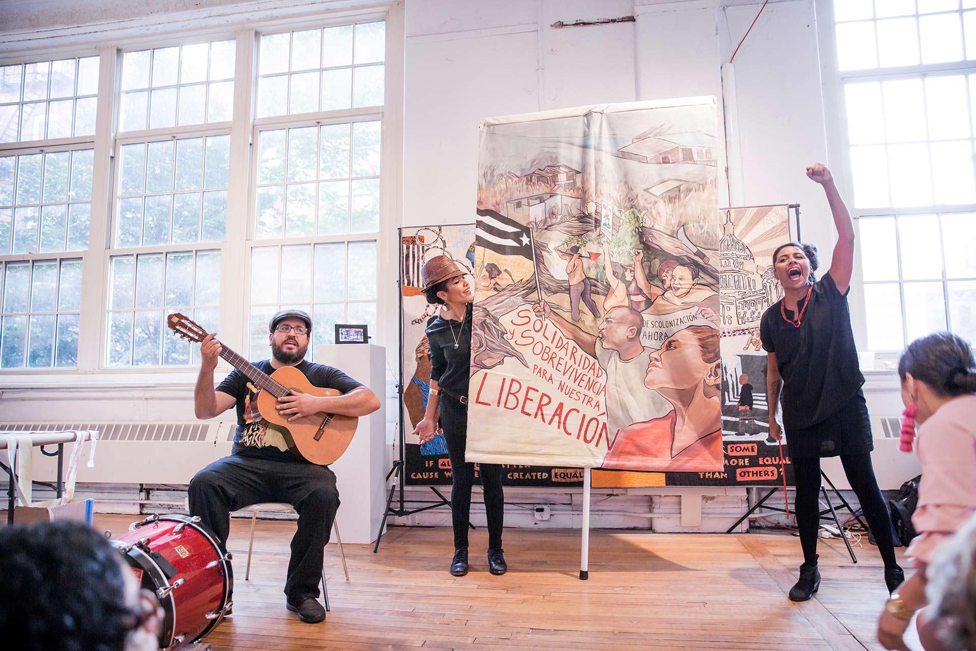 Three performers stand in front of a colorful banner about liberation inside a bright room; one plays guitar, one holds the banner, and one raises a fist while speaking passionately to an audience.