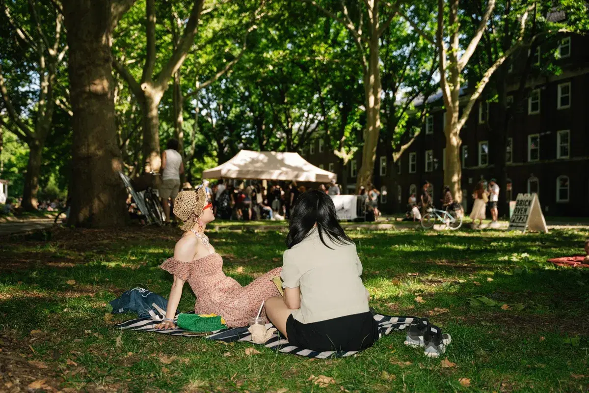 Two people sit on the grass with drinks and a picnic blanket 