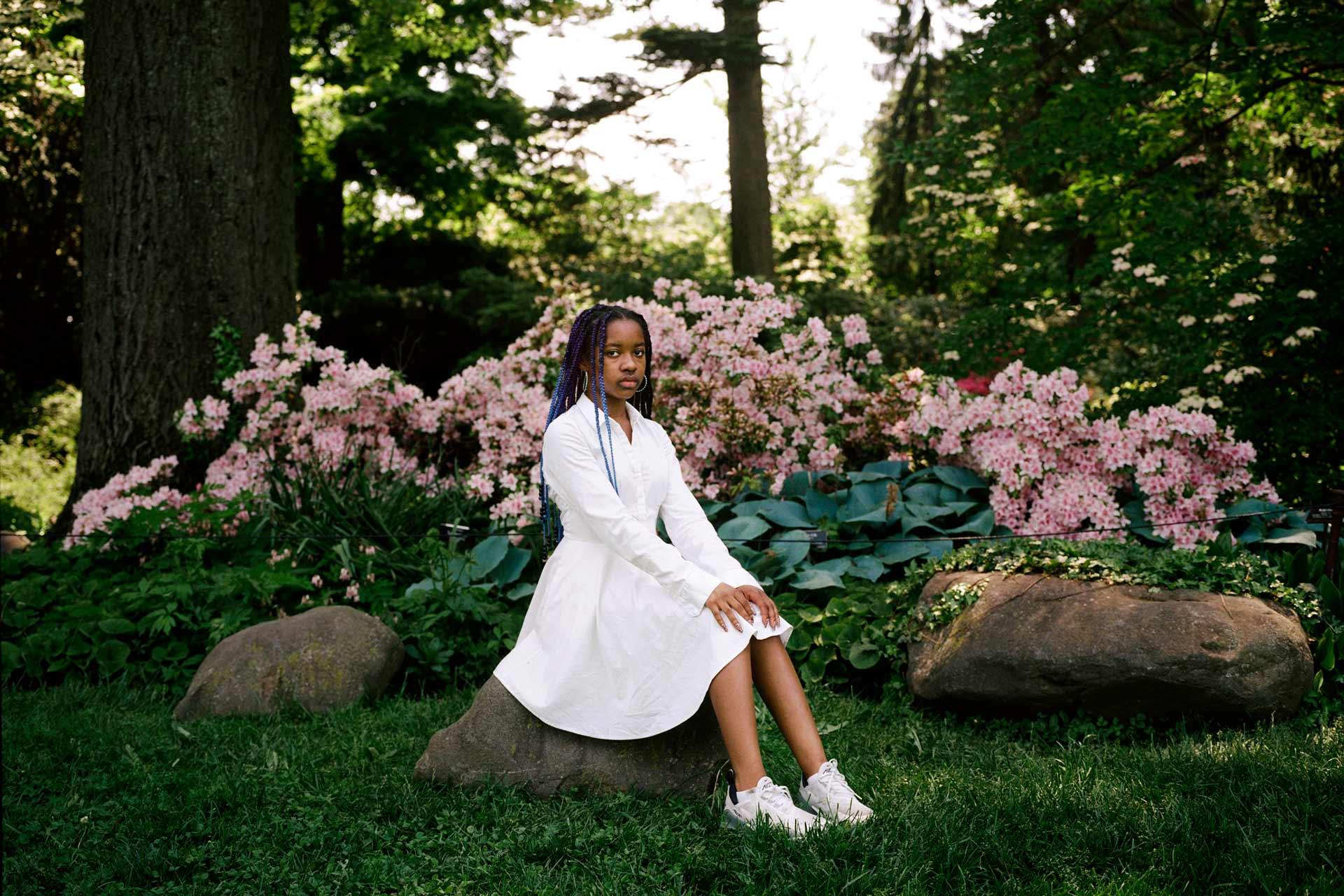 A portrait of a person sitting on a rock in a garden with flowers in the background. 