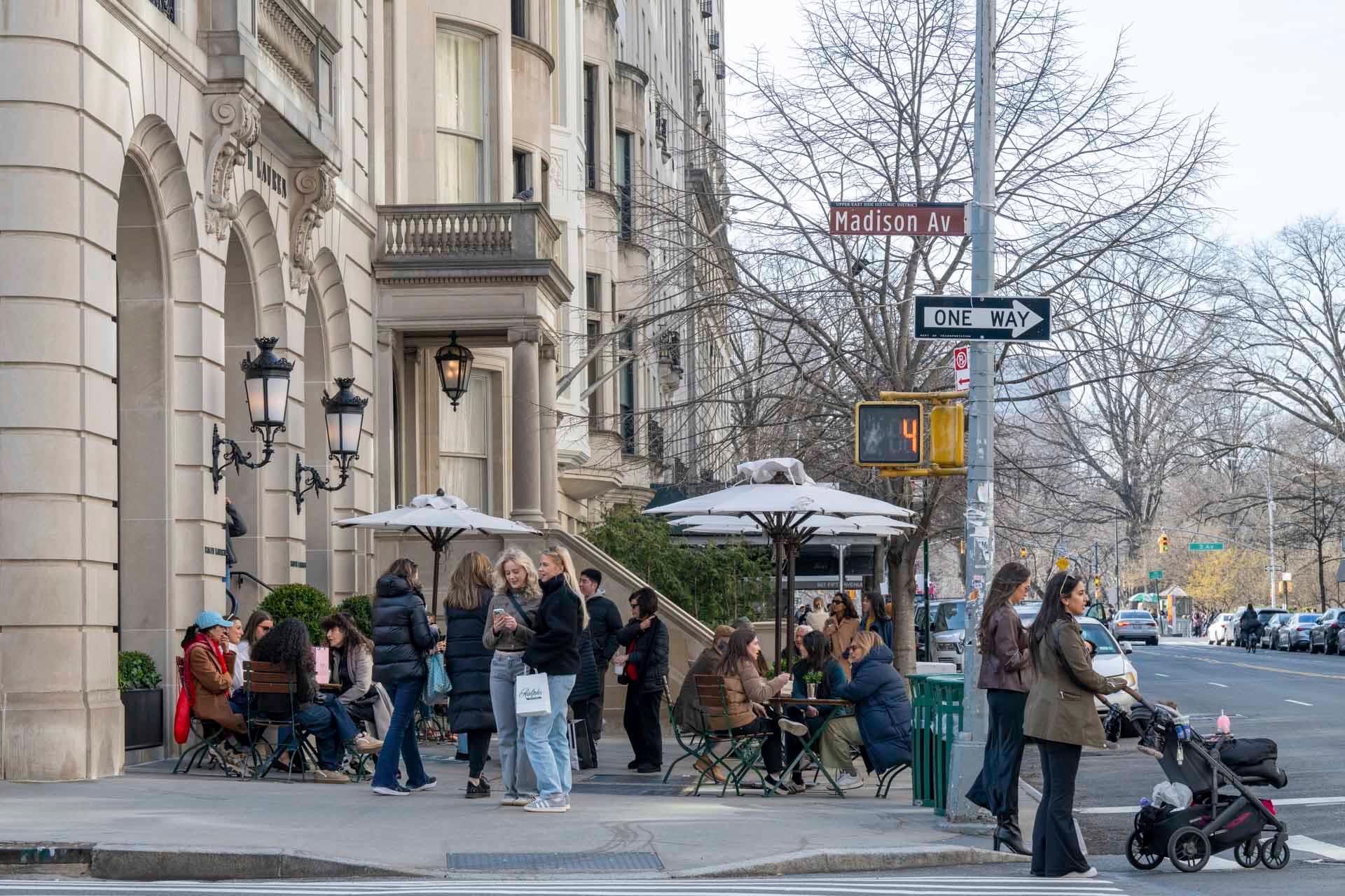 People gather outside a café on a busy city street corner. Many are sitting at tables under umbrellas, while others stand and chat. A street sign reads "Madison Av." Trees and a stroller-pushing person are visible in the background.