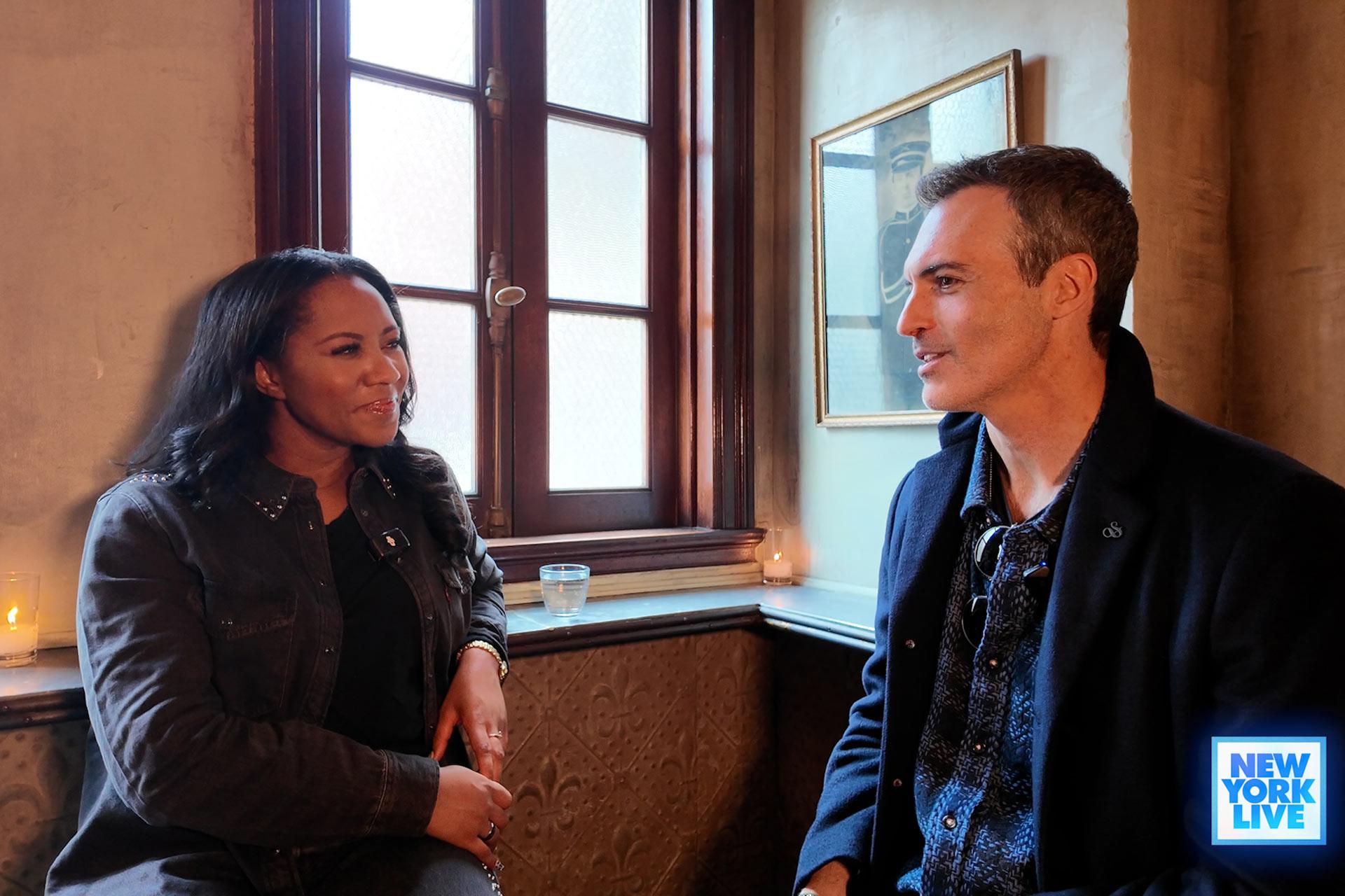 Two people sit and talk by a window in a cozy room with candles. The woman smiles and faces the man, who is attentively listening. A “New York Live” logo is in the bottom right corner.