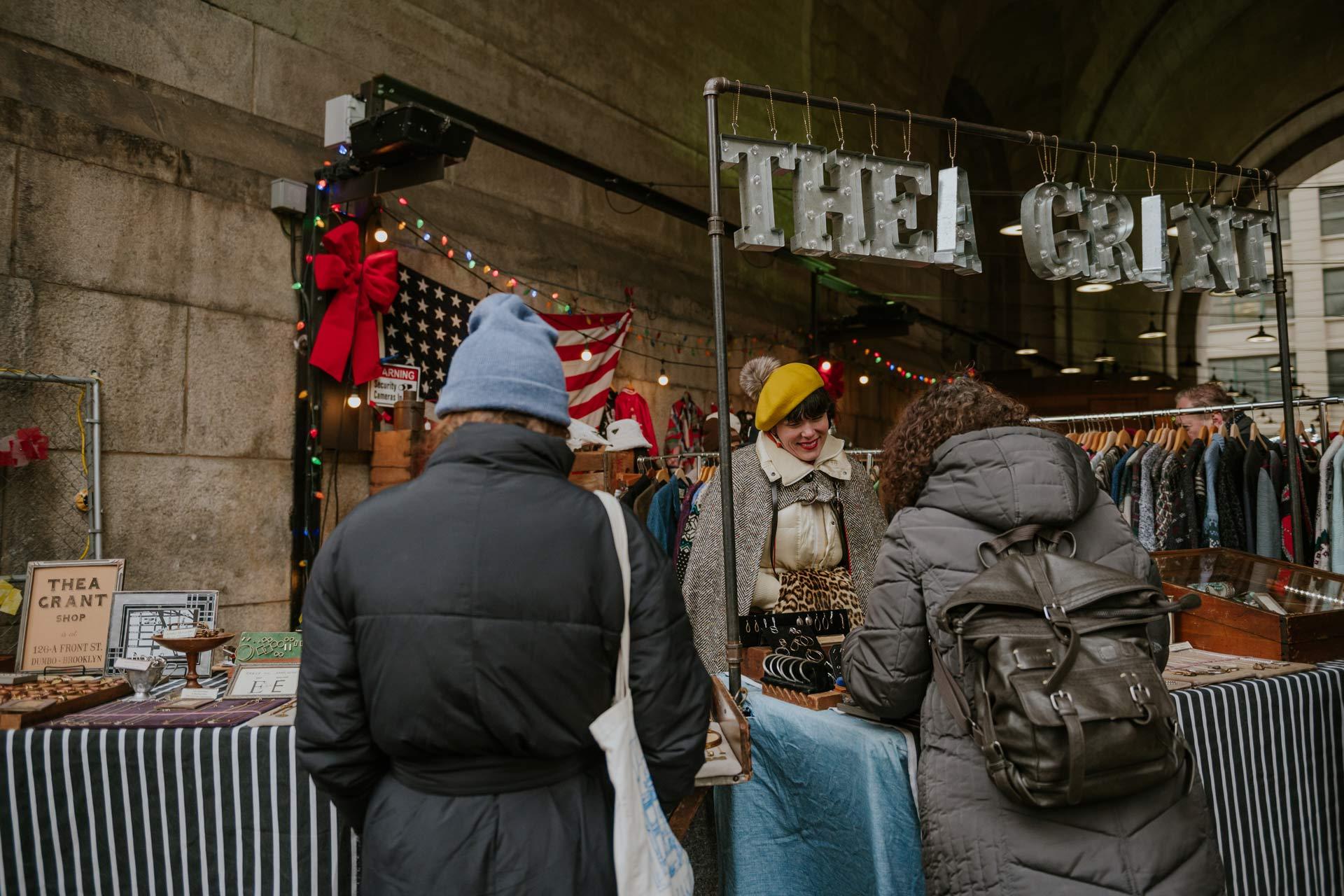 Two people in warm coats browse a stall at an outdoor market. The vendor smiles, wearing a yellow hat. The stall, decorated with festive lights and an American flag, displays various items. A sign above reads "THEA GRANT.