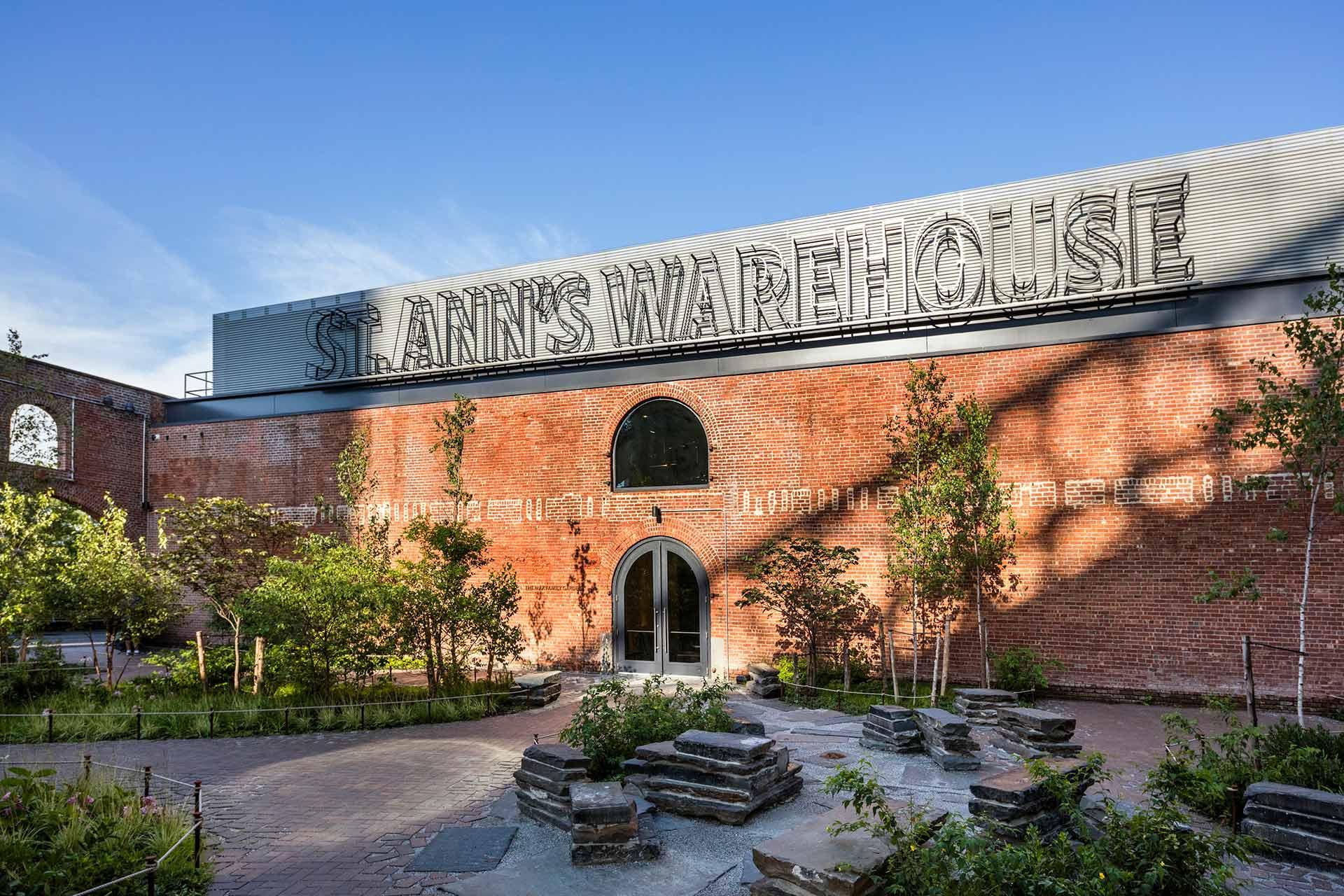 A brick building with large arched windows and a sign reading "ST. ANN'S WAREHOUSE" on the roof, surrounded by greenery and a landscaped garden under a clear blue sky.