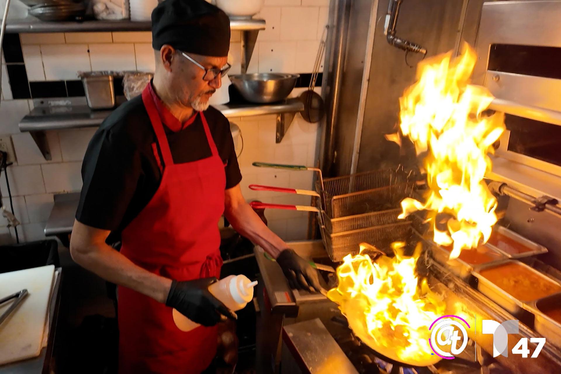 A chef in a red apron and black hat pours oil into a flaming pan in a commercial kitchen, creating a burst of fire, with kitchen equipment and utensils visible in the background.