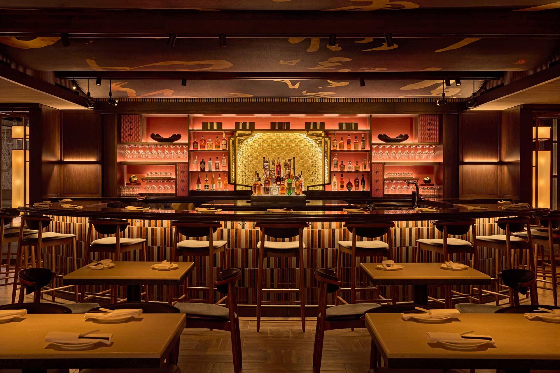 A stylish, warmly lit bar with wooden tables and chairs, a curved bar counter, and shelves of bottles behind. Soft amber lighting creates a cozy, inviting atmosphere with modern and elegant décor.