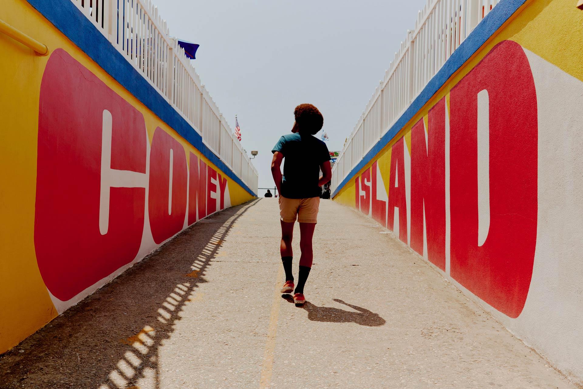 A person with short hair walks up a ramp between colorful walls painted with large red letters spelling "Coney Island," heading toward a bright, sunny sky.