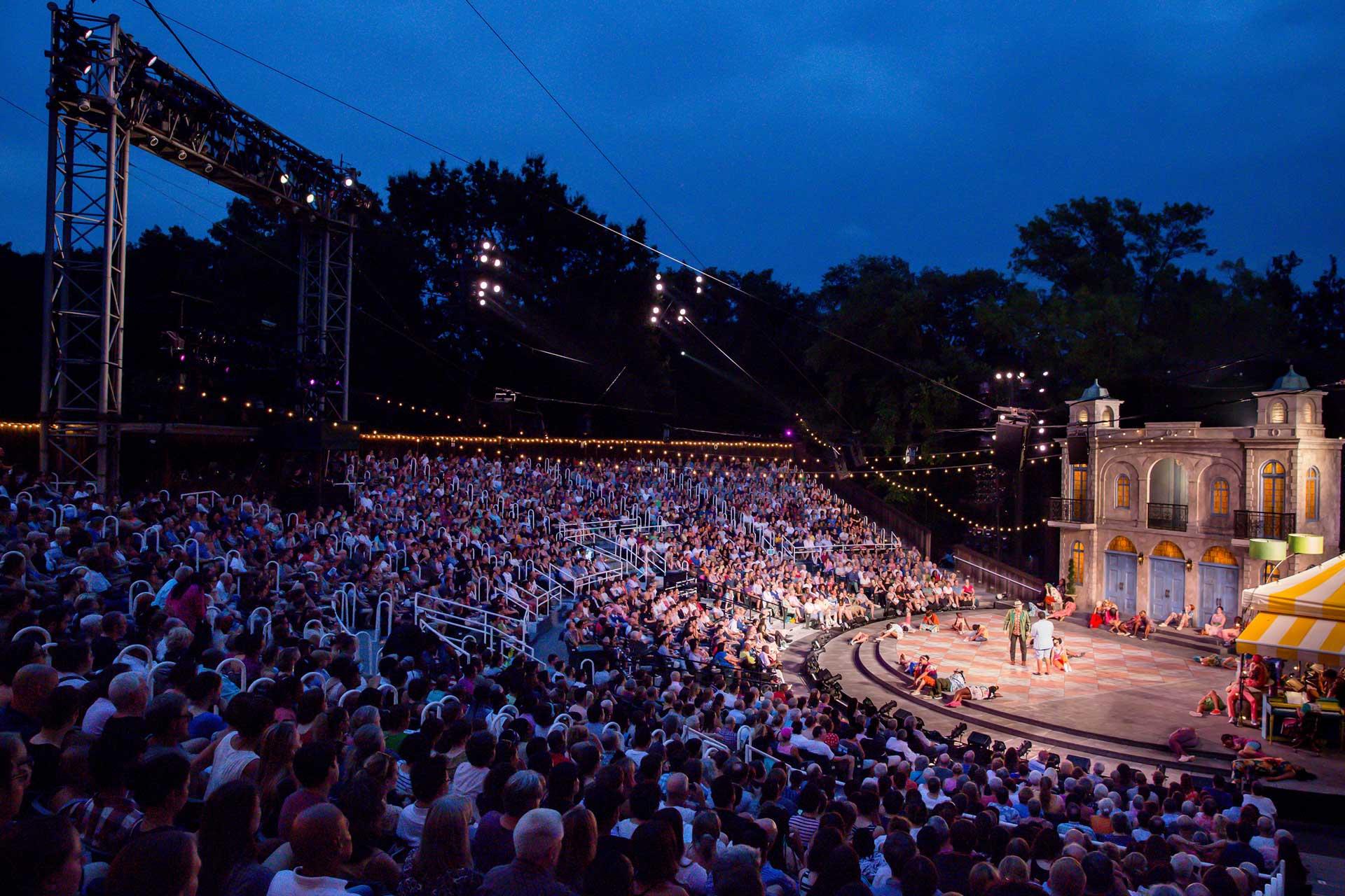 A large crowd watches an outdoor theater performance at dusk, with a brightly lit stage featuring several actors and a detailed set resembling buildings. Trees and stage lights surround the open-air venue.