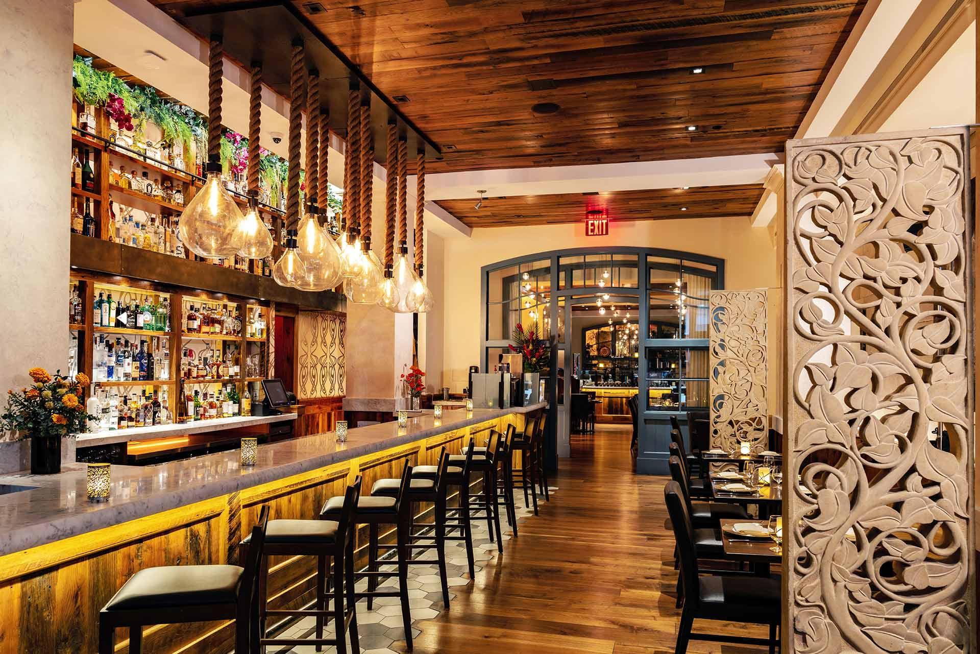 Stylish restaurant interior with a long bar lined with high chairs, hanging glass pendant lights, wooden floors and ceiling, decorative screen panels, and shelves of bottles behind the bar.