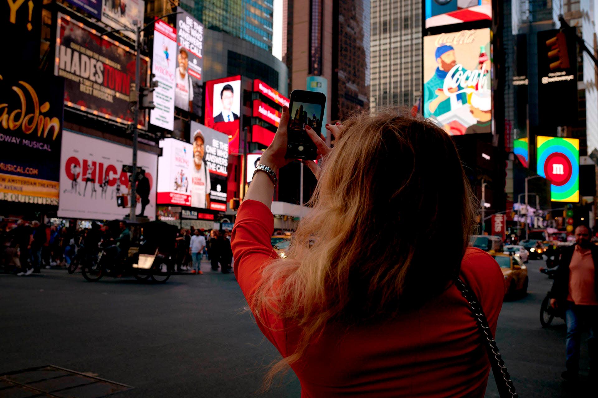 A person with long hair and an orange top takes a smartphone photo in Times Square, which is filled with bright billboards and advertisements.