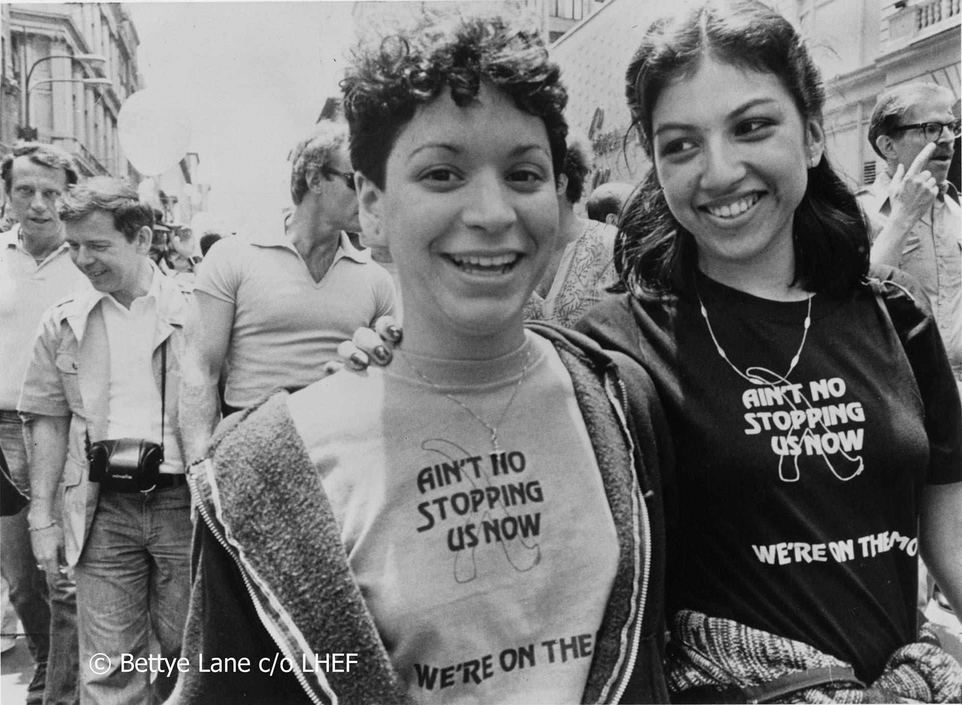 Two smiling young women at a street event wear matching “Ain’t No Stopping Us Now, We’re On The Move” shirts. People walk behind them on a sunny day. One woman has curly short hair; the other has long hair in braids.