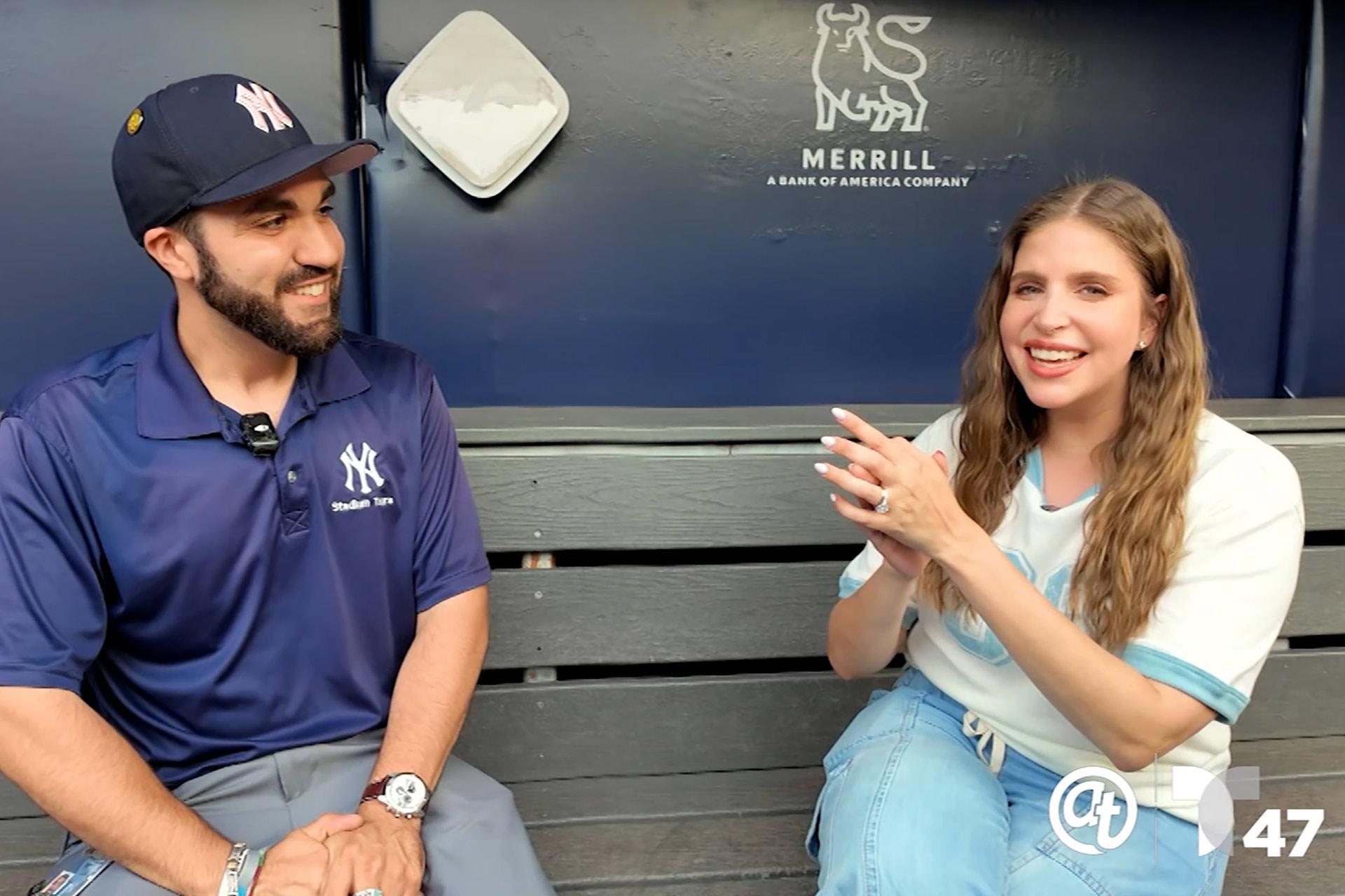 A man in a Yankees cap and uniform sits on a bench, smiling at a woman in a white and blue shirt who is clapping her hands. They appear to be having a friendly conversation in front of a Merrill sign.