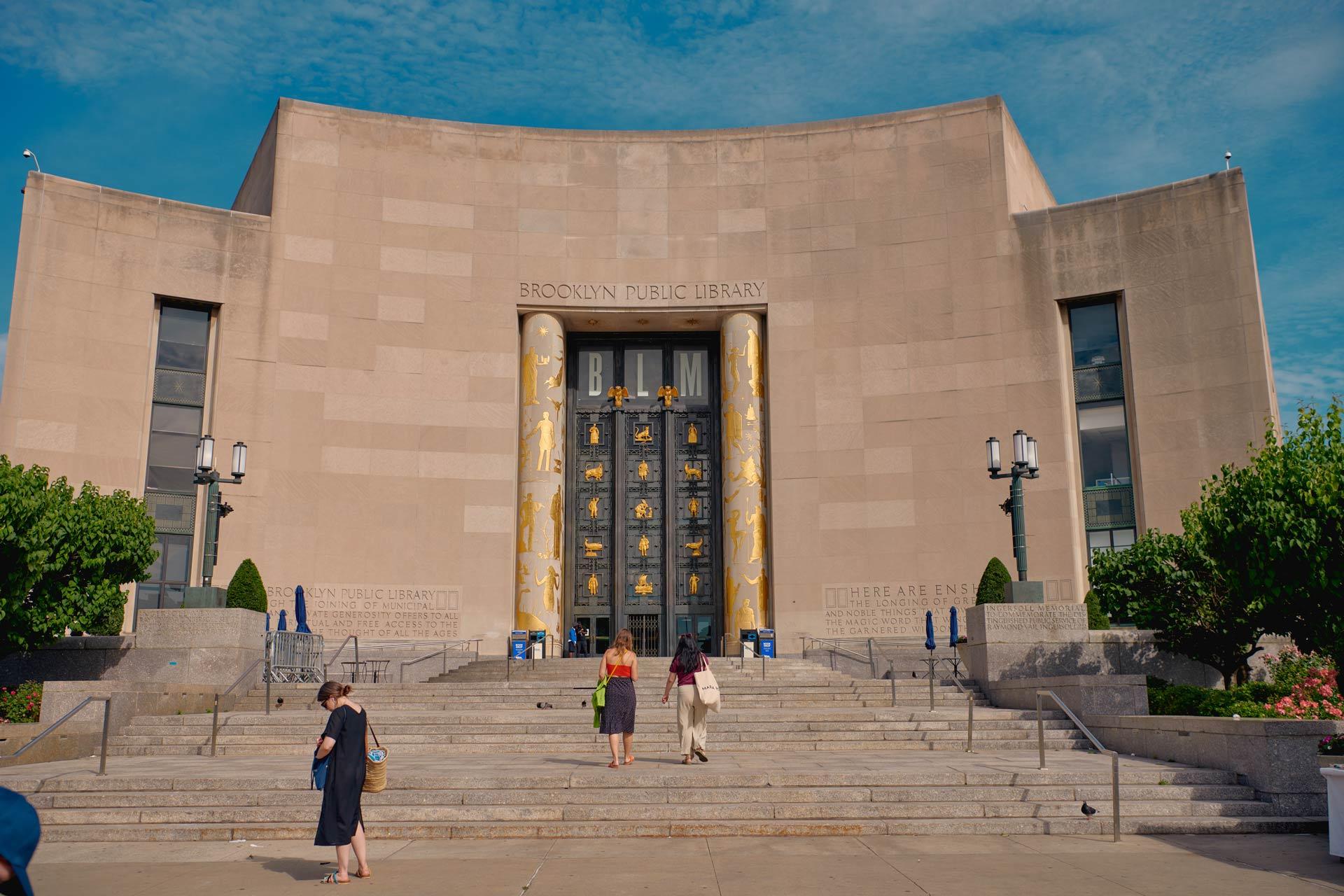 The image shows the main entrance of the Brooklyn Public Library. The grand building has a large, ornate door flanked by gold detailing and two women ascending the stairs. There are green bushes and lanterns on either side of the steps.
