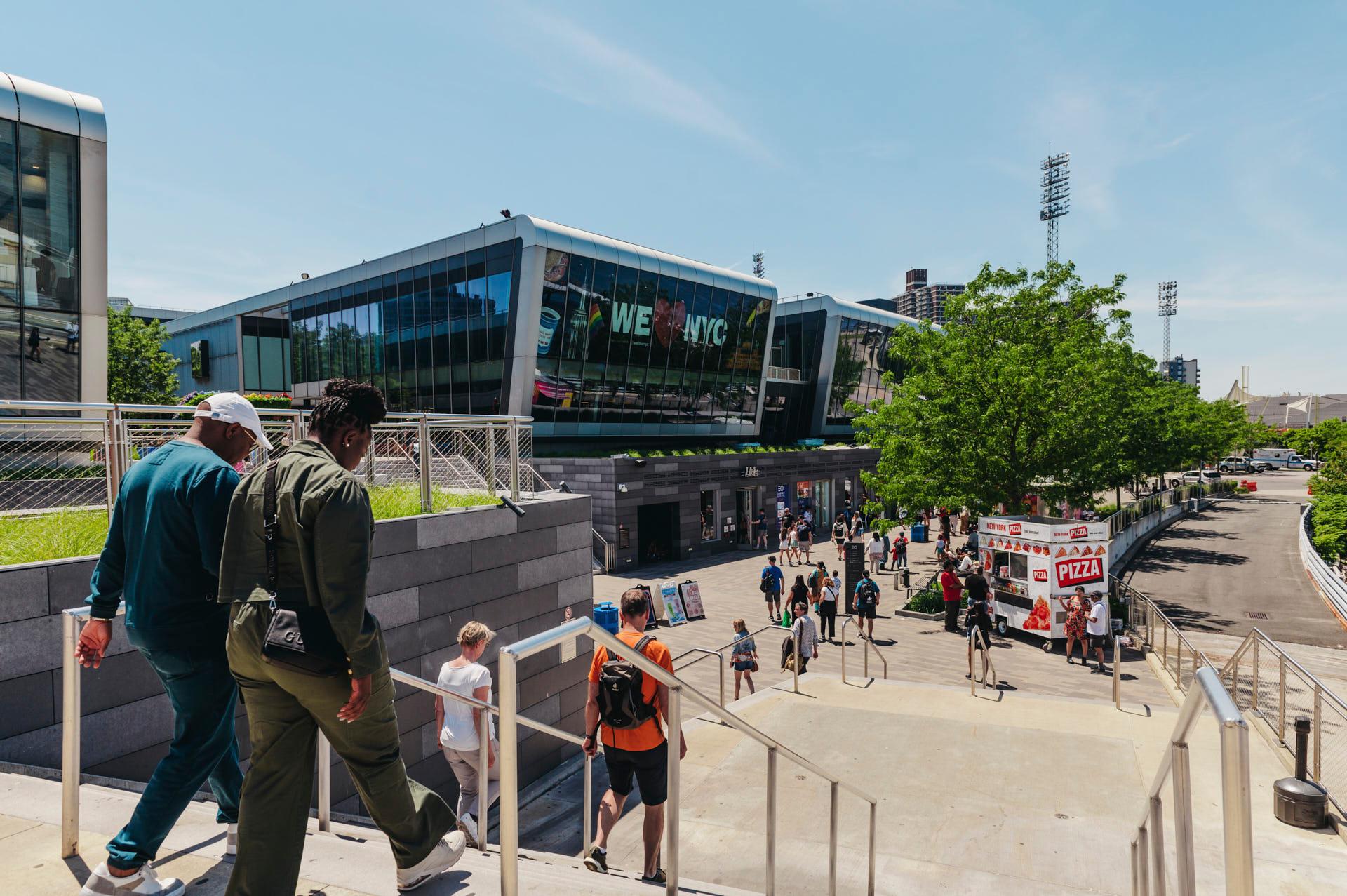 People walk down a staircase towards a modern building with glass windows on a sunny day. A pizza stand is visible to the right, and trees are scattered around the area. The building has "WE NYC" displayed on it.