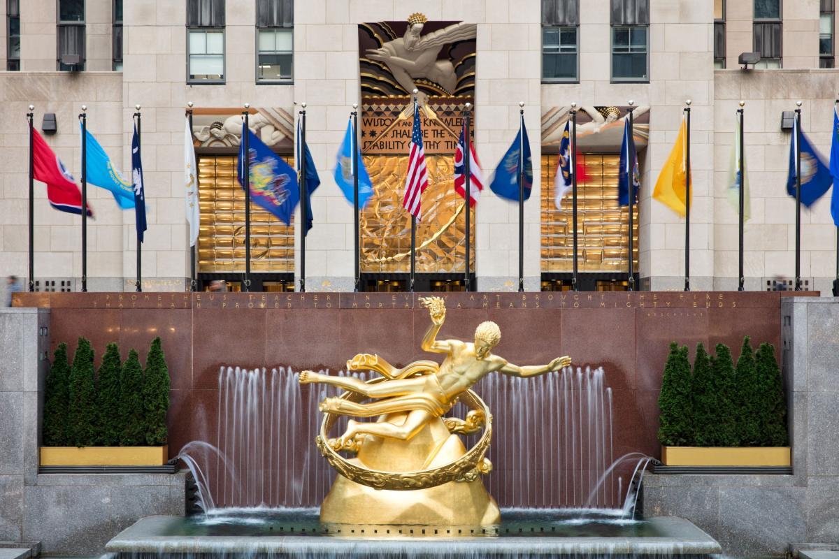 A golden statue of Prometheus stands in front of a fountain at Rockefeller Center, with national and state flags displayed above and a building entrance in the background.