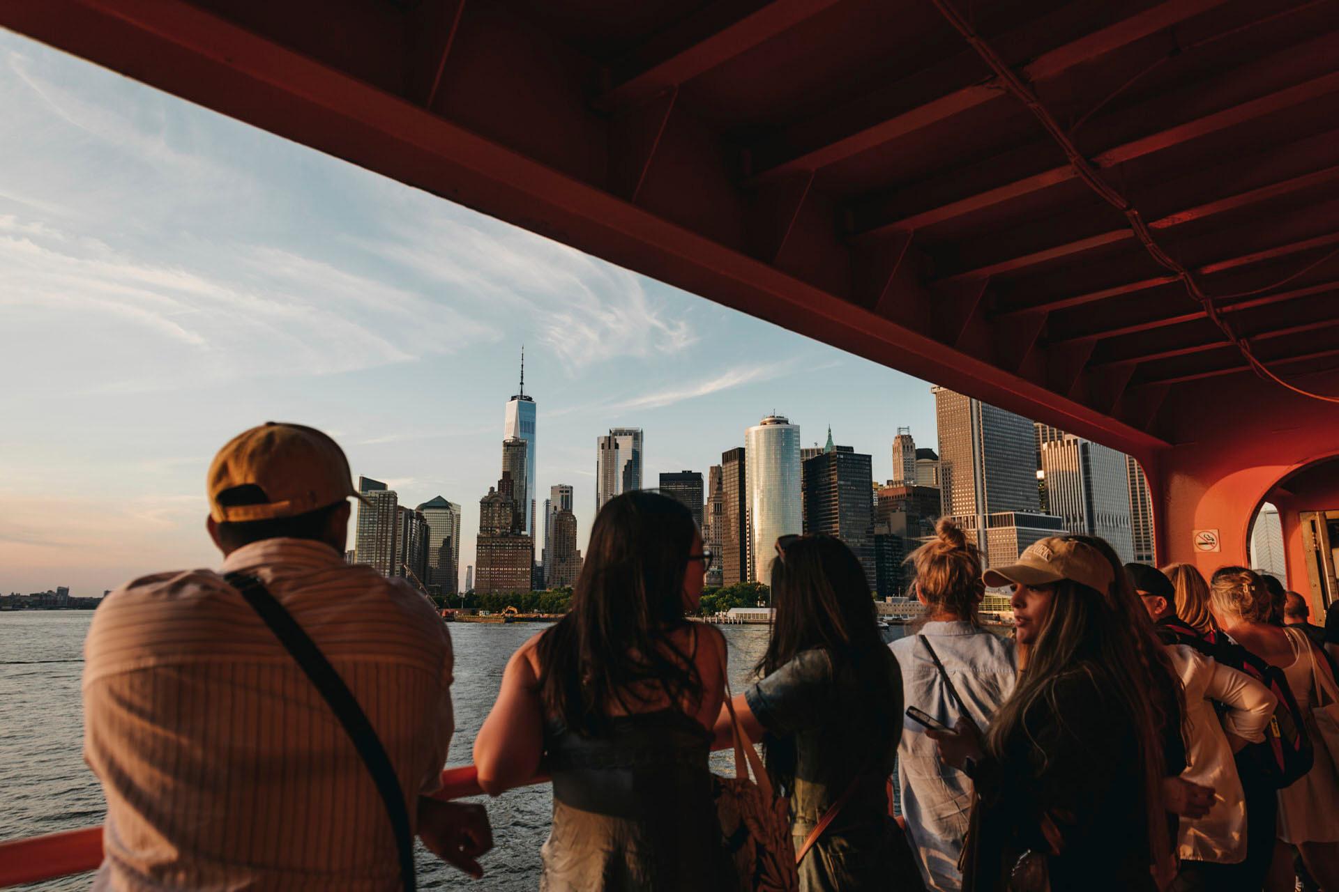 People stand on a ferry, looking at the New York City skyline featuring tall buildings and the distant view of One World Trade Center. The scene captures a warm, late afternoon light.