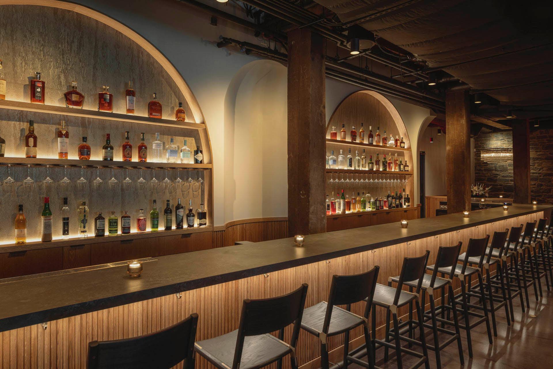 A modern bar interior with a long, wooden counter lined with stools. Shelves behind the bar display an array of liquor bottles. The space is warmly lit, creating a cozy ambiance.