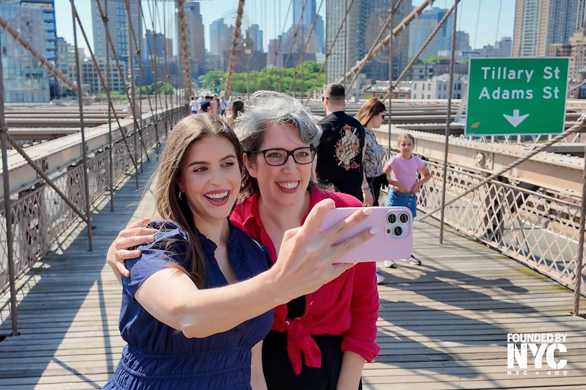 Two women smiling and taking a selfie together on the Brooklyn Bridge, with the New York City skyline and a green street sign reading "Tillary St Adams St" visible in the background. Other people are walking nearby.